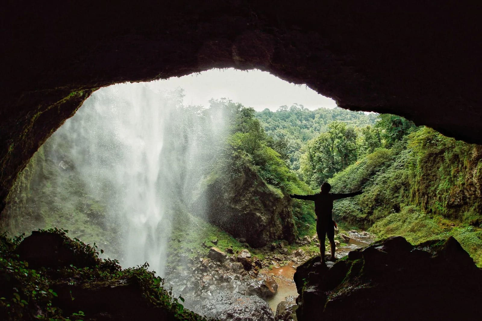 Grande cascade vue depuis une grotte, avec un randonneur en silhouette dans la végétation