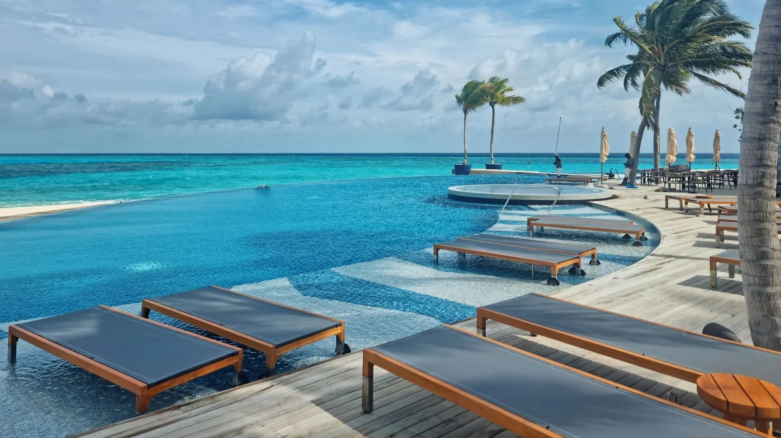 Piscine à débordement d'un hôtel face à un lagon turquoise, avec transats en bois et palmiers